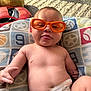 Jaxon joined the competition — help win amazing prizes! baby, child, glasses, orange_glasses, diaper, tongue_out, playful, cute, indoor, toy_car, stuffed_animal, pillow, patterned_pillow, carpet, infant, expression, face, skin, hand, relaxed