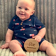 Colsen is registered to the contest to win money with this photo: baby, infant, smiling, sitting, onesie, american_flag, wooden_sign, six_months, blanket, texture, cute, child, person, happy, portrait, indoor, soft_lighting, skin, feet, hand