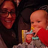 baby, blanket, car_seat, casual, child, closeup, face, glasses, happy, holding, indoor, infant, pacifier, person, smile, striped_blanket, toy, warm_lighting, woman, wooden_wall