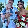 baby, celebration, daylight, family, fashion, flower_bouquet, formal_wear, greenery, happy, holding_baby, outdoor, person, portrait, red_bow_tie, red_dress, smiling, trees, white_suit, young_man, young_woman