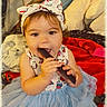 Sterling is registered to the contest to win money with this photo: baby, child, infant, portrait, sitting, smiling, bow, dress, tutu, blue, bed, blanket, pillow, indoors, cozy, remote_control, chewing, playful, cute, colorful