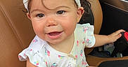 Sterling joined the competition — help win amazing prizes! baby, child, infant, sitting, car, car_interior, leather_seat, headband, bow, dress, patterned_clothing, smiling, happy, portrait, toy, colorful_toy, barefoot, indoors, vehicle, playful