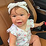 baby, child, infant, sitting, car, car_interior, leather_seat, headband, bow, dress, patterned_clothing, smiling, happy, portrait, toy, colorful_toy, barefoot, indoors, vehicle, playful