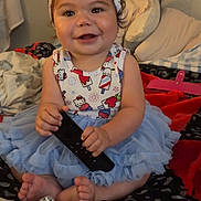 Sterling is registered to the contest to win money with this photo: baby, child, toddler, smiling, happy, portrait, indoor, sitting, bed, bedroom, headband, bow, dress, tutu, remote_control, blanket, pillows, colorful, artwork, barefoot