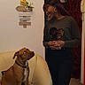 armchair, brown_dog, casual_clothing, collar, curious, curtain, dog, doorway, earrings, floor, home, indoor, interaction, long_hair, photograph, seated, smiling, standing, wall_decor, woman
