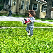 Legnadiel is registered to the contest to win money with this photo: activity, casual_clothing, child, curious, curly_hair, daytime, footwear, grass, greenery, house, lawn, outdoor, person, playing, side_view, street, toddler, tree, volleyball, young_child