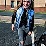 girl, child, smile, jeans, ripped_jeans, vest, blue_vest, long_hair, outdoor, parking_lot, sidewalk, building, brick_wall, asphalt, casual_clothing, person, young, happy, sunlight, walking