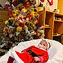 baby, christmas_tree, christmas_decorations, red_clothing, pacifier, headband, cushion, holiday_stockings, indoor, festive, holiday, smiling, person, furniture, wooden_shelf, lights, ornaments, celebration, cozy, child