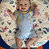 baby, smiling, infant, blue_onesie, dinosaur_pattern, cushion, colorful, carpet, playful, happy, child, person, floor, feet, socks, cute, indoors, portrait, lying_down, bright