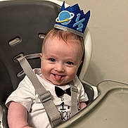 Declan is registered to the contest to win money with this photo: baby, high_chair, birthday, crown, blue_crown, tongue_out, white_shirt, bow_tie, smiling, infant, child, straps, seat, face, happy, cheeky, indoor, person, cute, celebration