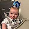 baby, high_chair, birthday, crown, blue_crown, tongue_out, white_shirt, bow_tie, smiling, infant, child, straps, seat, face, happy, cheeky, indoor, person, cute, celebration