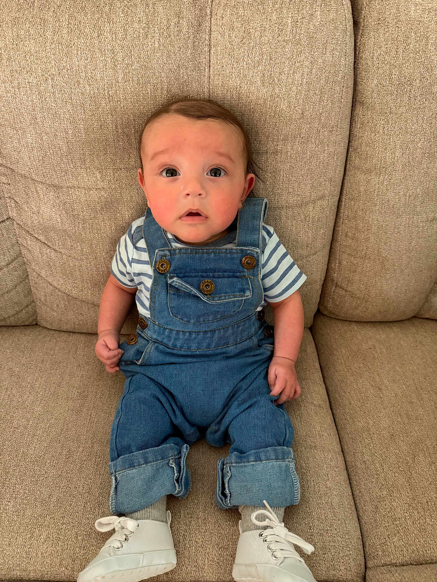 Malachi is registered to the contest to win money with this photo: baby, infant, denim_overalls, white_shoes, striped_shirt, couch, beige, seated, curious_expression, cute, child, indoors, portrait, small_hands, fashion, footwear, casual_clothing, young_child, face, eyes