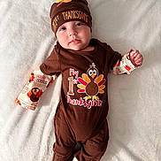 Malachi joined the competition — help win amazing prizes! baby, infant, thanksgiving, holiday, brown_clothing, hat, onesie, turkey_graphic, white_blanket, cute, smiling, portrait, child, feet, hand, lying_down, festive, soft_texture, celebration, indoors