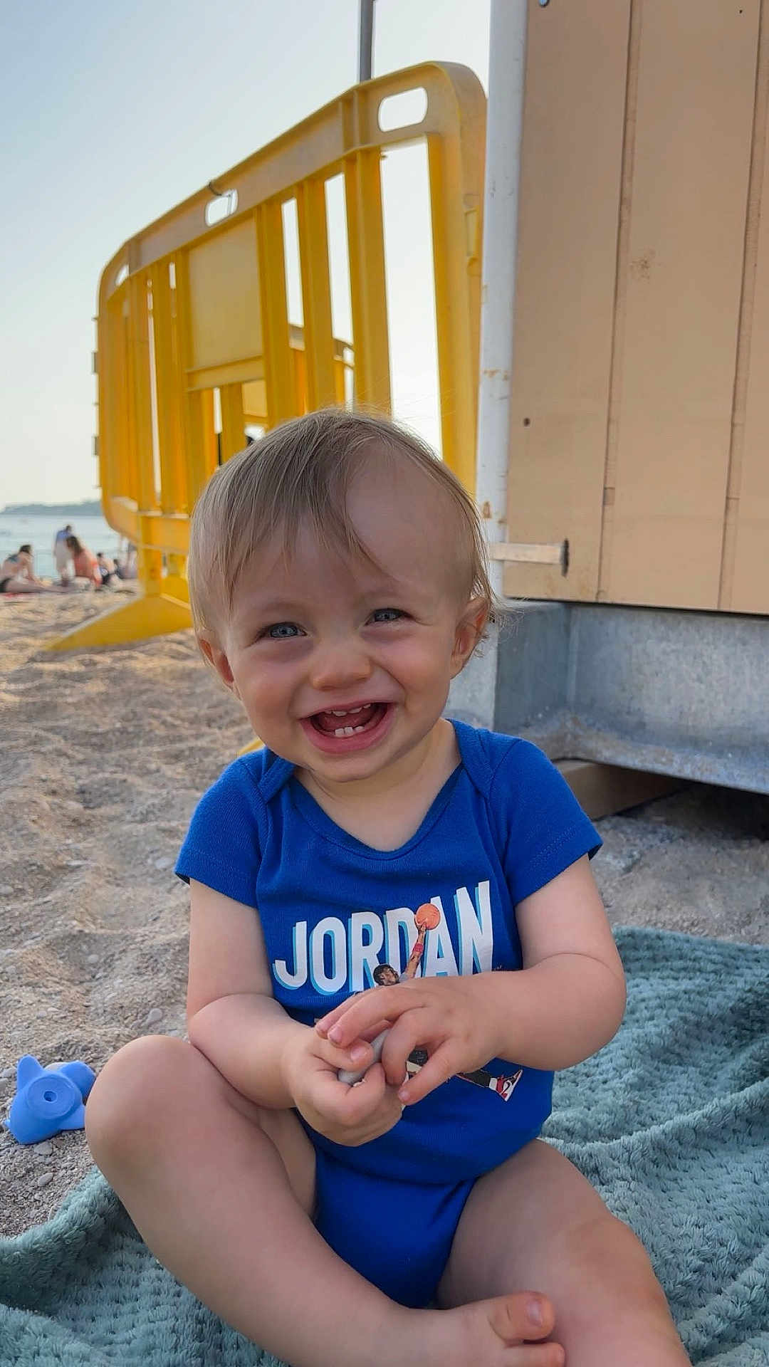 Eyden a rejoint le concours — aidez-le/la à gagner de superbes lots ! toddler, child, smiling, beach, sand, blue_clothing, onesie, happy, outdoor, sunny, blanket, toy, barrier, yellow, person, portrait, sitting, cute, daylight, casual