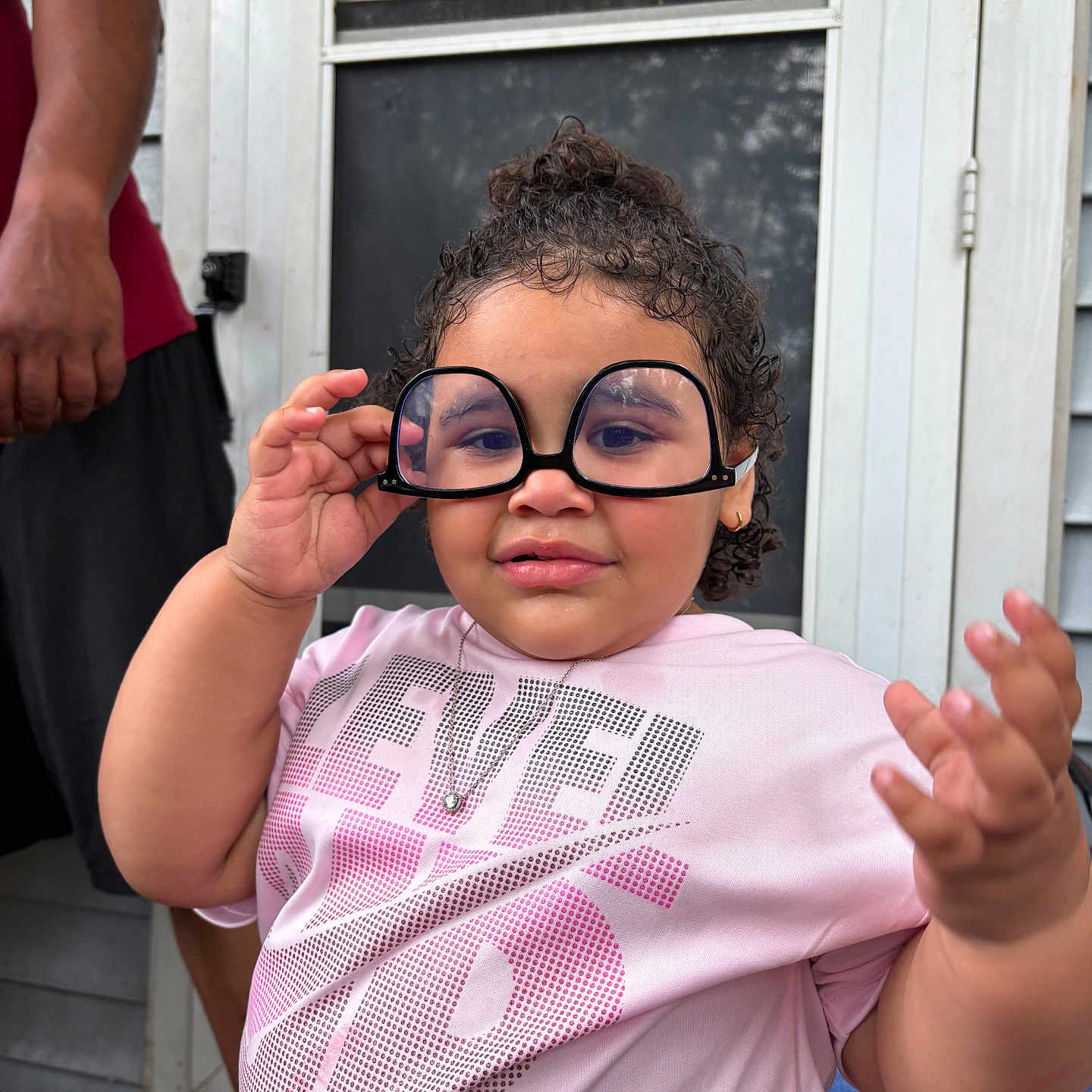 Lilth is registered to the contest to win money with this photo: adult_arm, casual_clothing, child, curly_hair, door, expression, funny_glasses, glasses, hand, home, necklace, outdoor, person, pink_shirt, playful, portrait, screen_door, smile, toddler