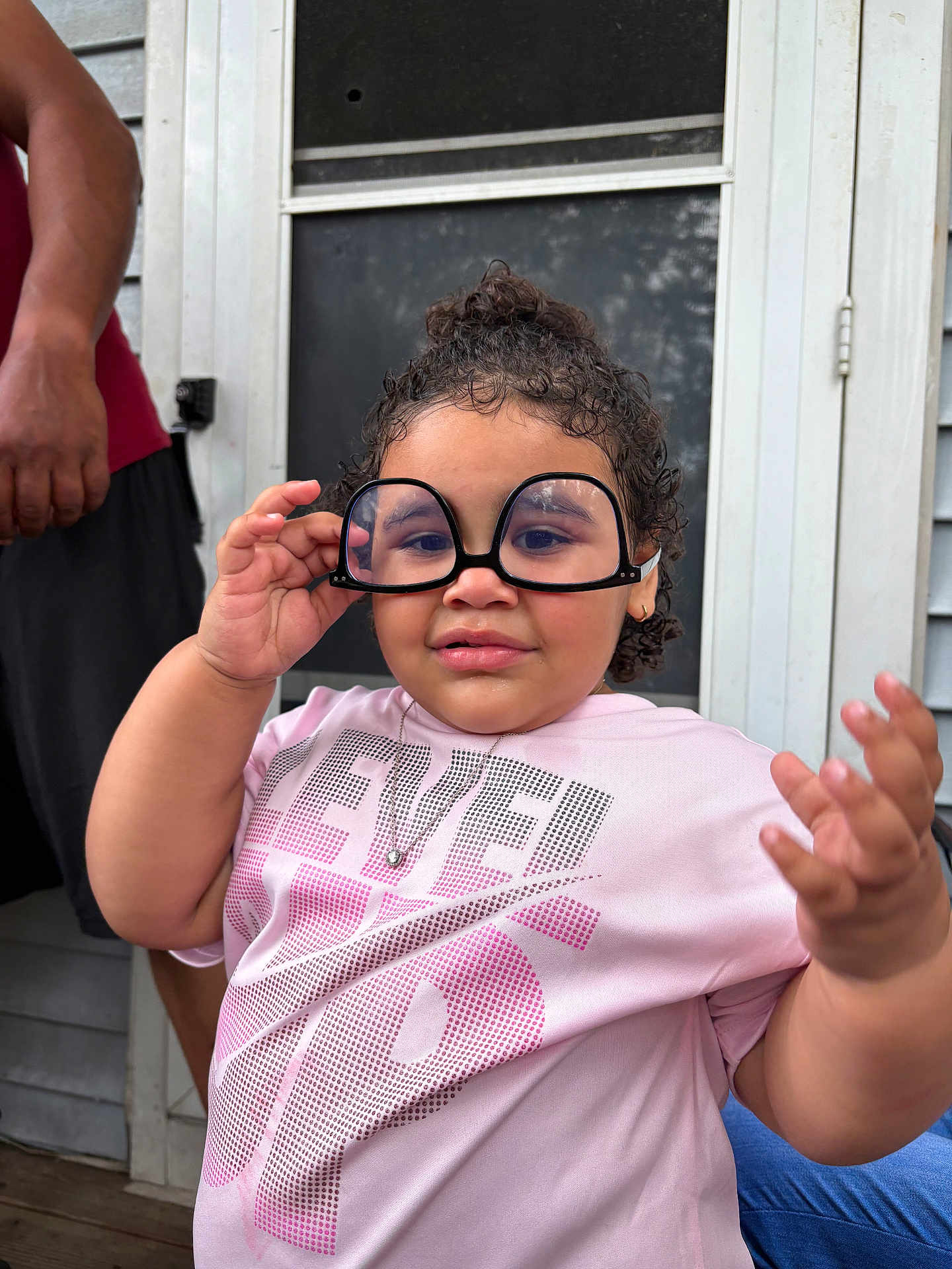 Lilth is registered to the contest to win money with this photo: adult_arm, casual_clothing, child, curly_hair, door, expression, funny_glasses, glasses, hand, home, necklace, outdoor, person, pink_shirt, playful, portrait, screen_door, smile, toddler