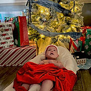 Harper joined the competition — help win amazing prizes! baby, christmas_tree, presents, red_blanket, red_booties, holiday, festive, ornaments, soft_cushion, wooden_floor, indoors, decorations, infant, holiday_lighting, wrapped_gifts, celebration, smiling_baby, cozy, christmas_hat, warm_lighting