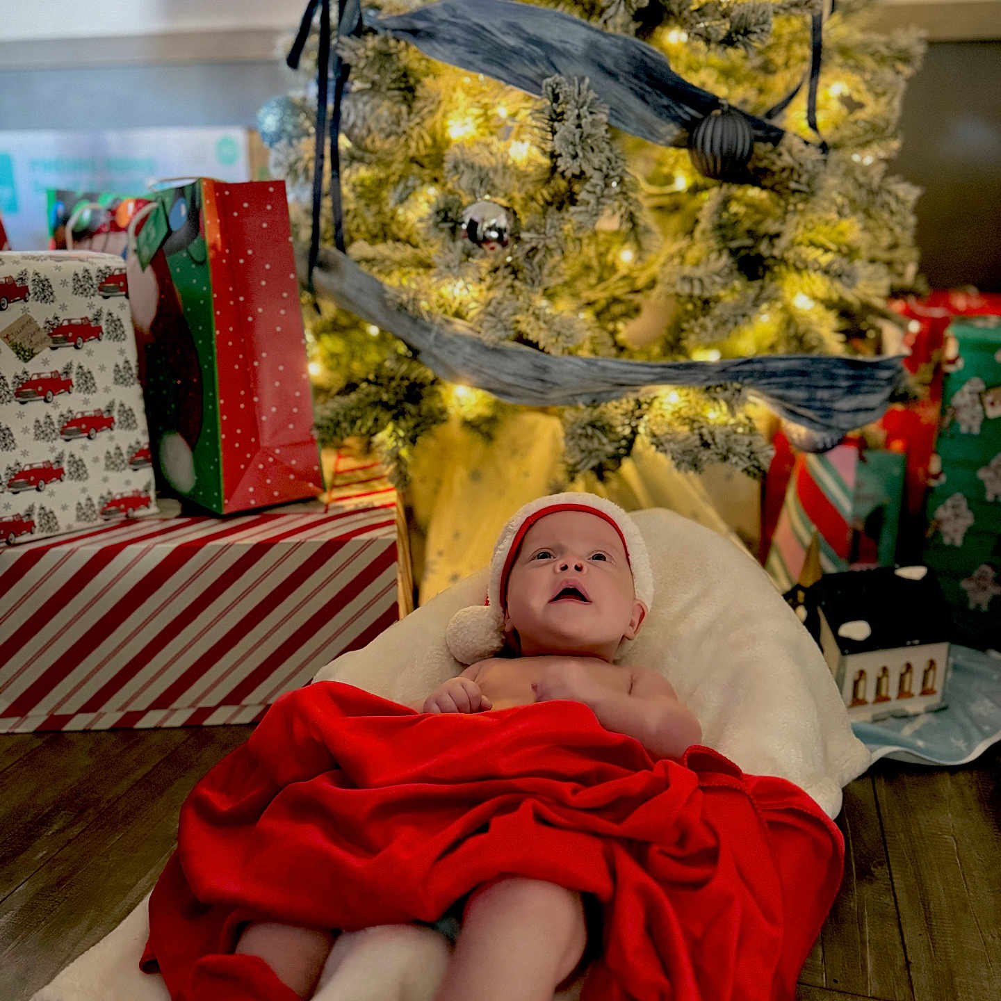 Harper joined the competition — help win amazing prizes! baby, celebration, christmas_hat, christmas_tree, cozy, decorations, festive, holiday, holiday_lighting, indoors, infant, ornaments, presents, red_blanket, red_booties, smiling_baby, soft_cushion, warm_lighting, wooden_floor, wrapped_gifts