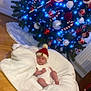 baby, blanket, christmas_tree, ornaments, blue_lights, wooden_floor, hat, red_hat, white_clothing, holiday, festive, decoration, indoors, infant, candy_cane, red_ornament, white_ornament, pine_tree, cozy, celebration