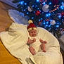 baby, infant, hat, blanket, christmas_tree, ornaments, lights, floor, holiday, festive, red, white, wood, decorations, indoor, smiling, cute, seasonal, cozy, celebration