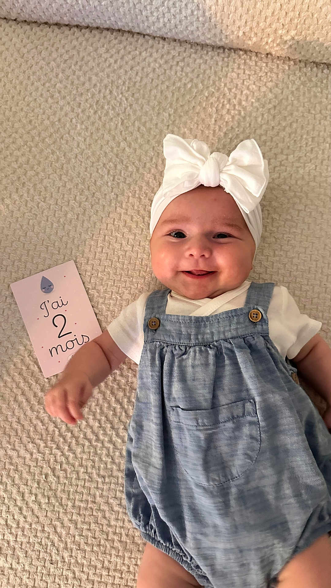 Victoire participe au concours pour gagner de l'argent avec cette photo : age_card, baby, blanket, blue_clothing, bow, card, child, closeup, cute, happy, headband, indoors, infant, lying_down, person, portrait, romper, smiling, soft_texture, white_shirt