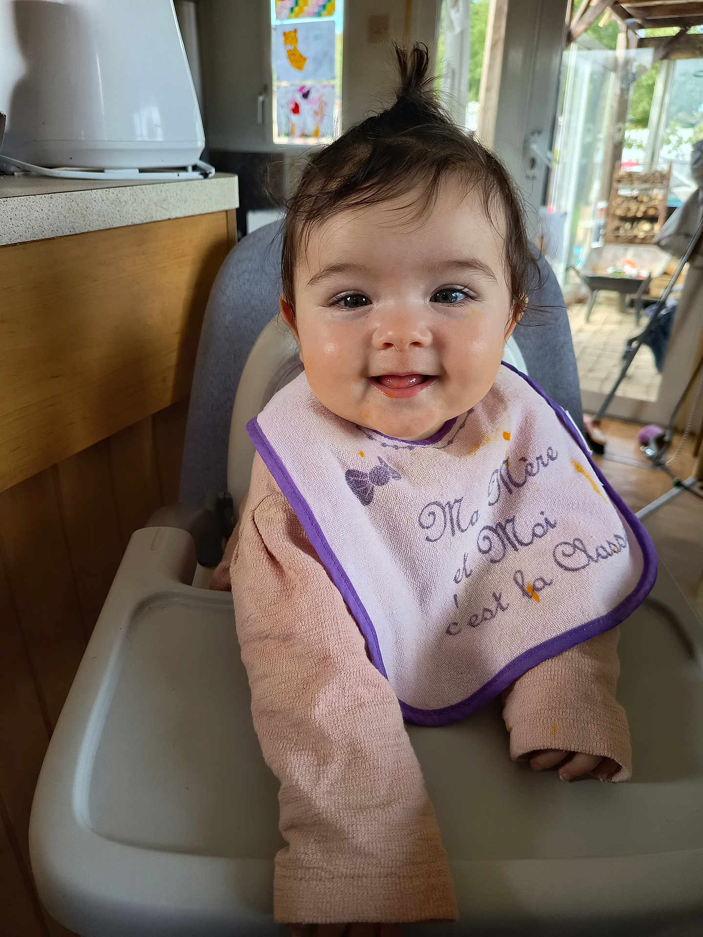 Anita participe au concours pour gagner de l'argent avec cette photo : baby, child, high_chair, bib, smile, indoors, kitchen, window, sunlight, hair, long_sleeve, face, happy, infant, person, cozy, messy_face, pink_clothing, wooden_cabinet, feeding