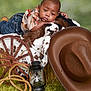 baby, child, jeans, shirt, cowhide_blanket, wooden_wheel, rope, lantern, cowboy_hat, green_background, fur, portrait, studio_photo, infant, resting, props, western_theme, cute, indoor, soft_texture