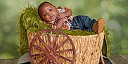 Ezekiel joined the competition — help win amazing prizes! baby, child, basket, wooden_wheel, green_background, jeans, shirt, brown_shoes, cowhide_cushion, floor, indoor, portrait, resting, cute, woven, furniture, fuzzy_blanket, infant, relaxed, decor