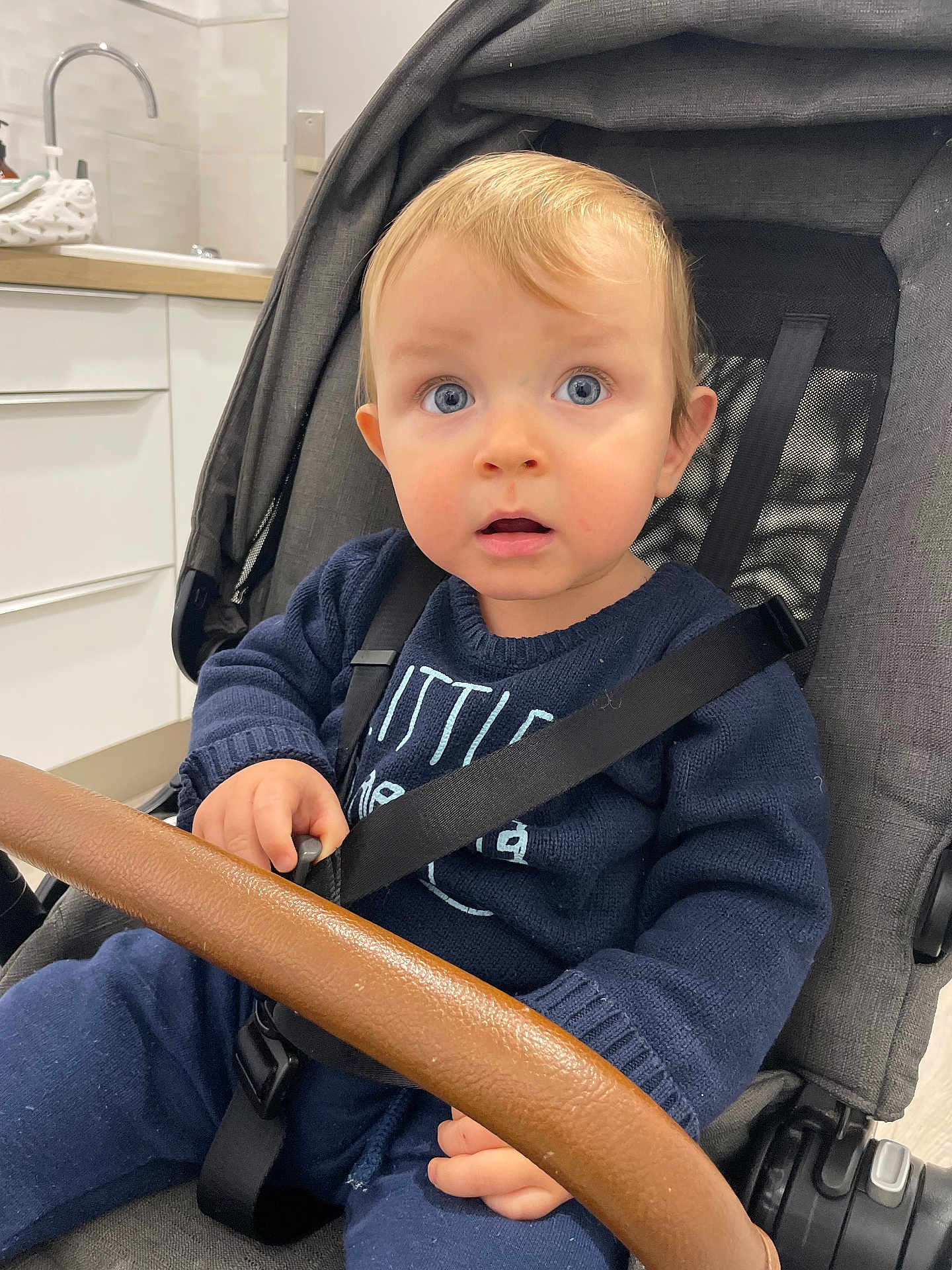 Lino participe au concours pour gagner de l'argent avec cette photo : baby, child, blue_eyes, stroller, seatbelt, navy_sweater, indoor, kitchen, sink, faucet, countertop, seat, hand, face, person, clothing, expression, curly_hair, young, sitting