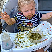 Lino participe au concours pour gagner de l'argent avec cette photo : toddler, baby_food, high_chair, messy, child, food, feeding, plate, bottle, clothing, face, expression, indoor, hair, seat, table, arm, hand, skin, curious