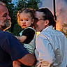 child, toddler, man, woman, outdoor, family, holding, smoke, trees, nature, evening, casual_clothing, glasses, beard, portrait, curious, greenery, light, people, backdrop