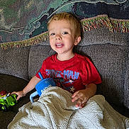 Gabriel is registered to the contest to win money with this photo: child, boy, red_shirt, spiderman, toy, toy_tractor, blanket, couch, sippy_cup, indoor, smiling, happy, face, person, furniture, home, relaxing, seated, blanket_texture, casual