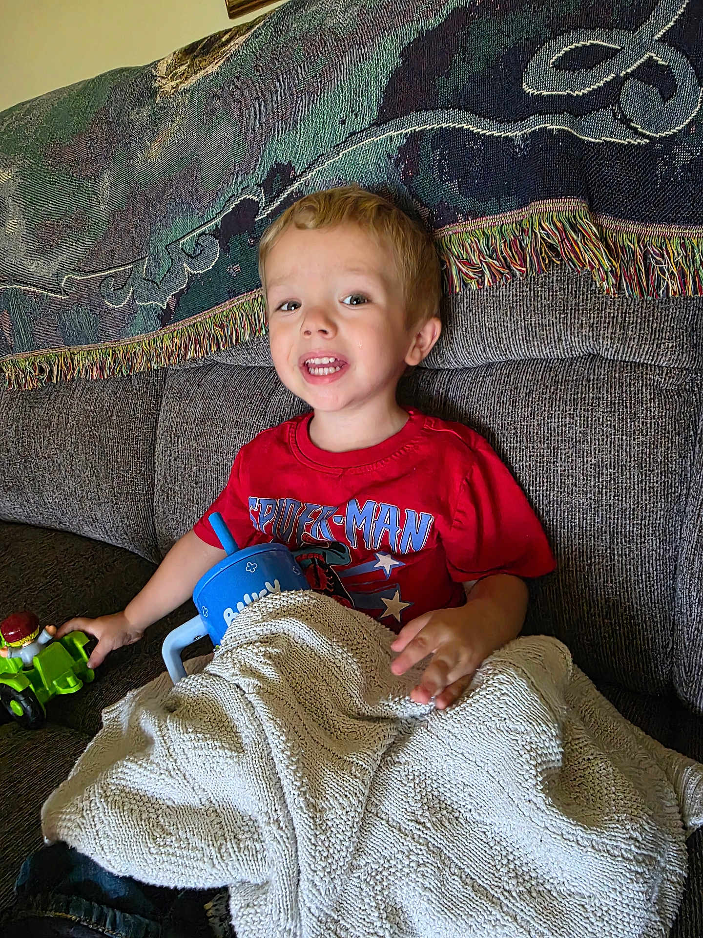 Gabriel is registered to the contest to win money with this photo: child, boy, red_shirt, spiderman, toy, toy_tractor, blanket, couch, sippy_cup, indoor, smiling, happy, face, person, furniture, home, relaxing, seated, blanket_texture, casual