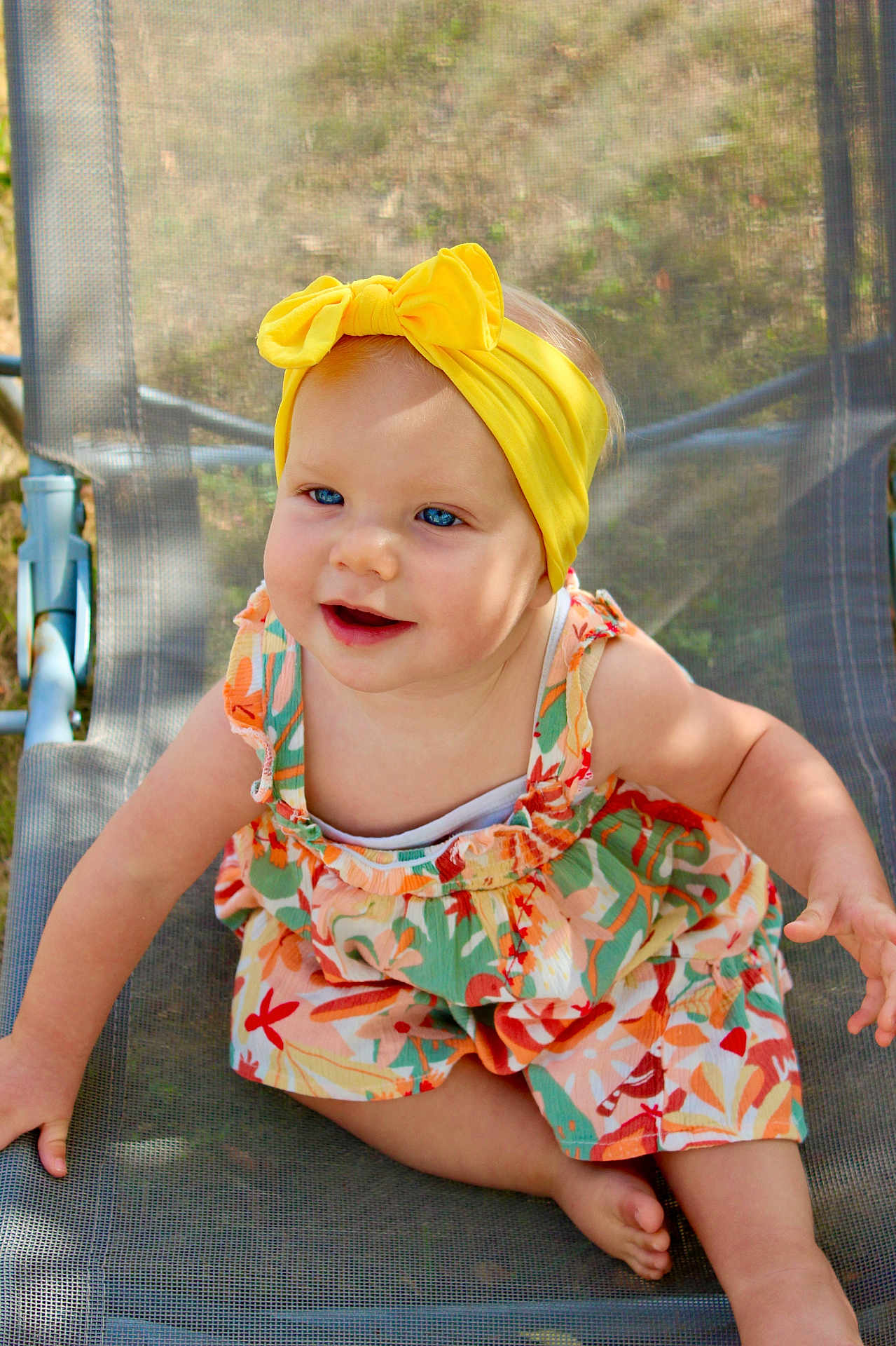 Cléo a rejoint le concours — aidez-le/la à gagner de superbes lots ! baby, child, girl, yellow_headband, blue_eyes, smiling, outdoor, sunlight, floral_dress, mesh_chair, barefoot, happy, cute, summer, sitting, portrait, one_person, infant, headwear, colorful_clothing