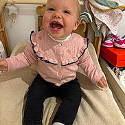 Cléo participe au concours pour gagner de l'argent avec cette photo : baby, child, smiling, pink_cardigan, black_pants, white_socks, indoor, cushioned_surface, happy, clothing, footwear, bib, baby_shoes, furniture, room, portrait, cute, joyful, person, sitting