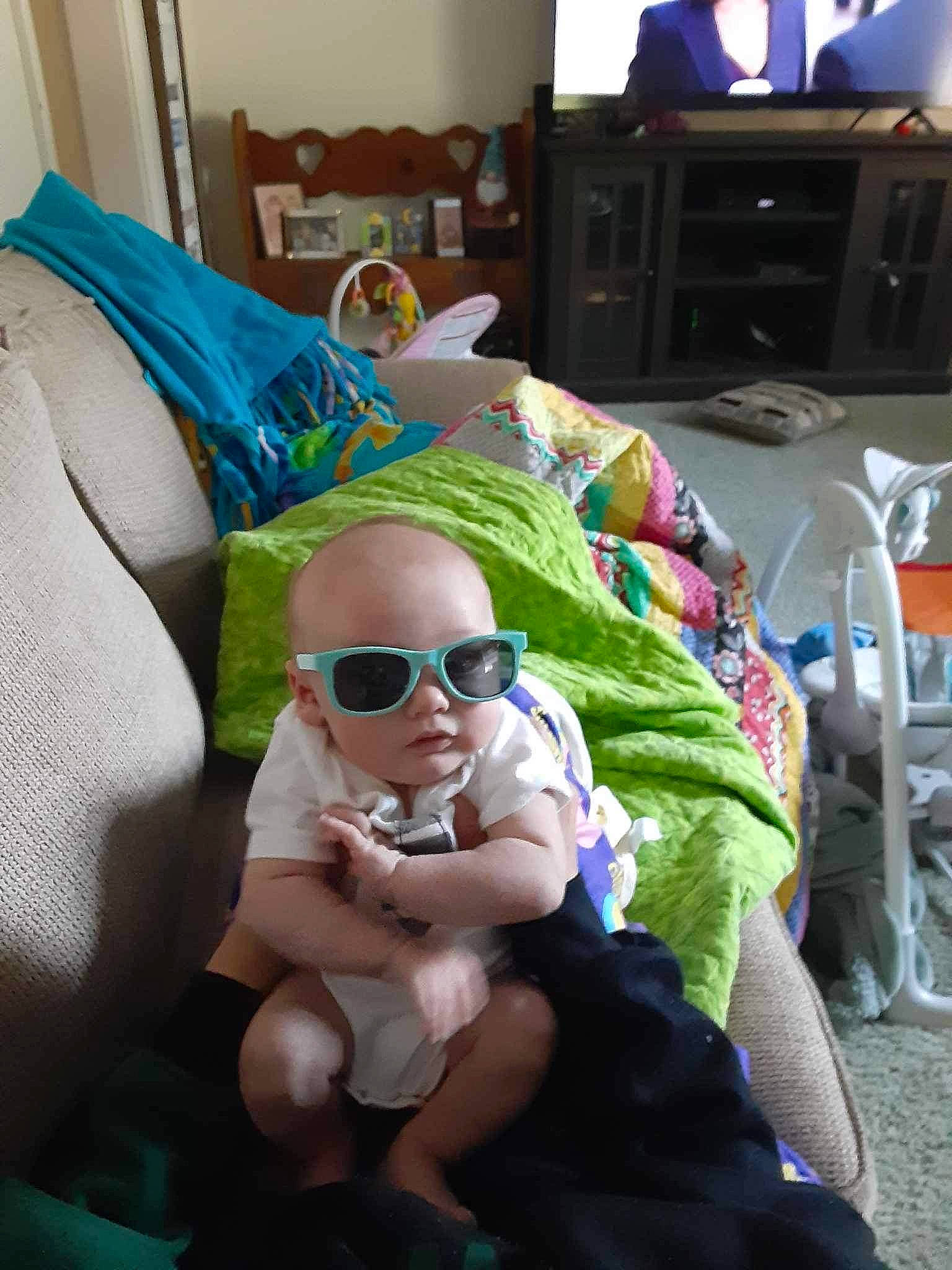 Cameron is registered to the contest to win money with this photo: baby, baby_products, comfort, event, eyewear, foot, fun, glasses, goggles, human_leg, lap, leisure, person, room, sitting, smile, sunglasses, television, thigh, toddler