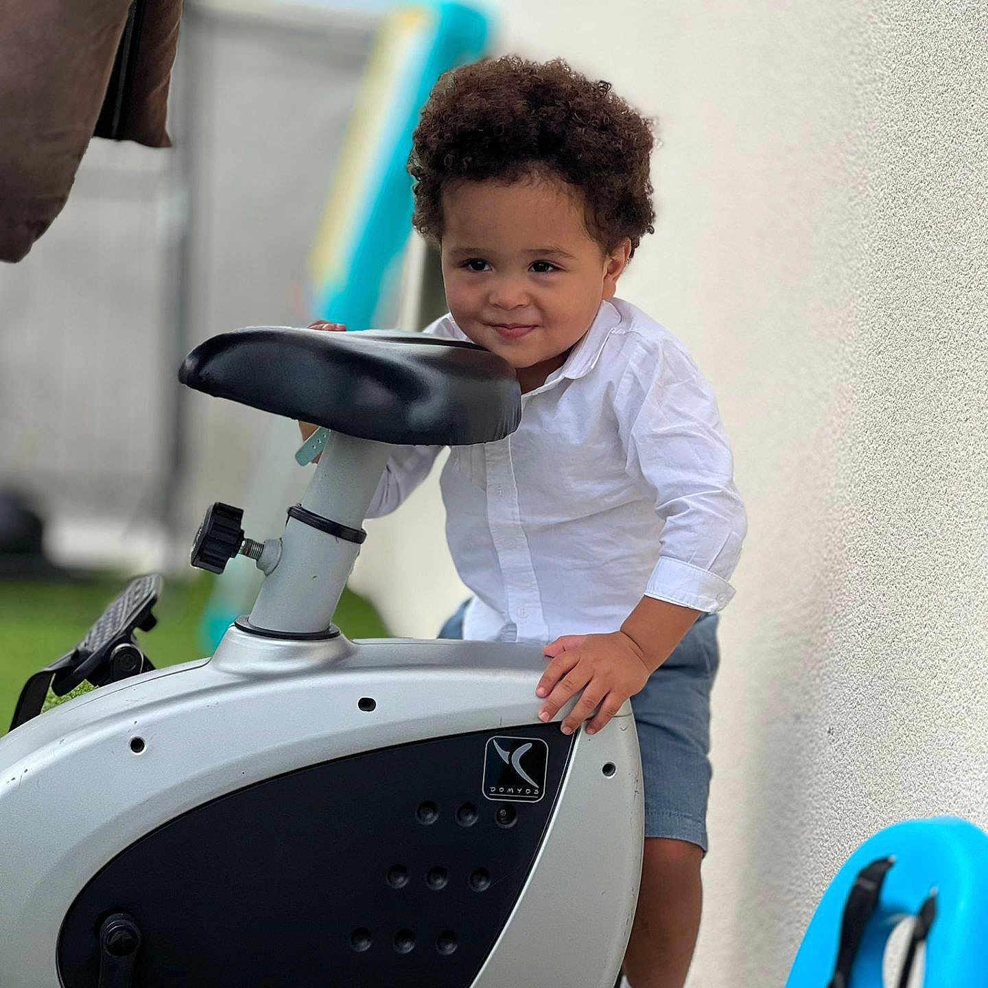 Heitor Benício participe au concours pour gagner de l'argent avec cette photo : blue_shorts, casual_clothing, child, curly_hair, daylight, exercise_bike, grass, happy, outdoor, person, playful, shoes, smiling, sport_equipment, standing, toddler, toy, wall, white_shirt, young_child