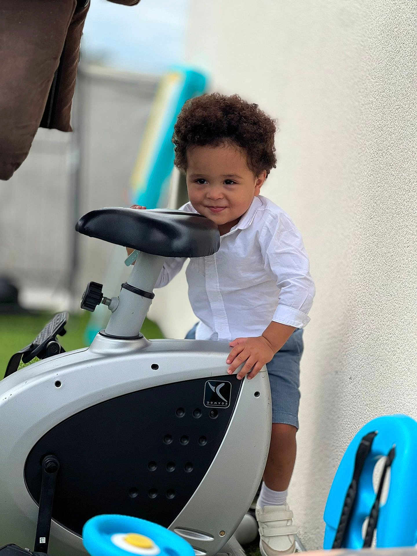 Heitor Benício participe au concours pour gagner de l'argent avec cette photo : toddler, child, curly_hair, white_shirt, blue_shorts, exercise_bike, outdoor, playful, smiling, person, toy, shoes, wall, grass, daylight, happy, young_child, standing, sport_equipment, casual_clothing