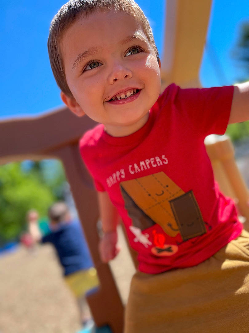 Diego is registered to the contest to win money with this photo: baby, child, city, finger, fun, gesture, grass, happy, joy, leisure, outdoor_play_equipment, person, recreation, sand, sitting, sky, smile, swing, t_shirt, thumb