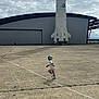 background, building, casual_clothing, child, cloudy, concrete, daytime, footwear, hangar, hat, large_structure, motion, open_space, outdoor, play, rocket, running, sky, space, toddler