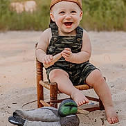Nicholas is registered to the contest to win money with this photo: toddler, child, smiling, cap, brown_cap, overalls, camouflage, wooden_chair, sand, toy, wooden_toy, duck_decoy, outdoor, nature, greenery, happy, barefoot, portrait, seat, summer