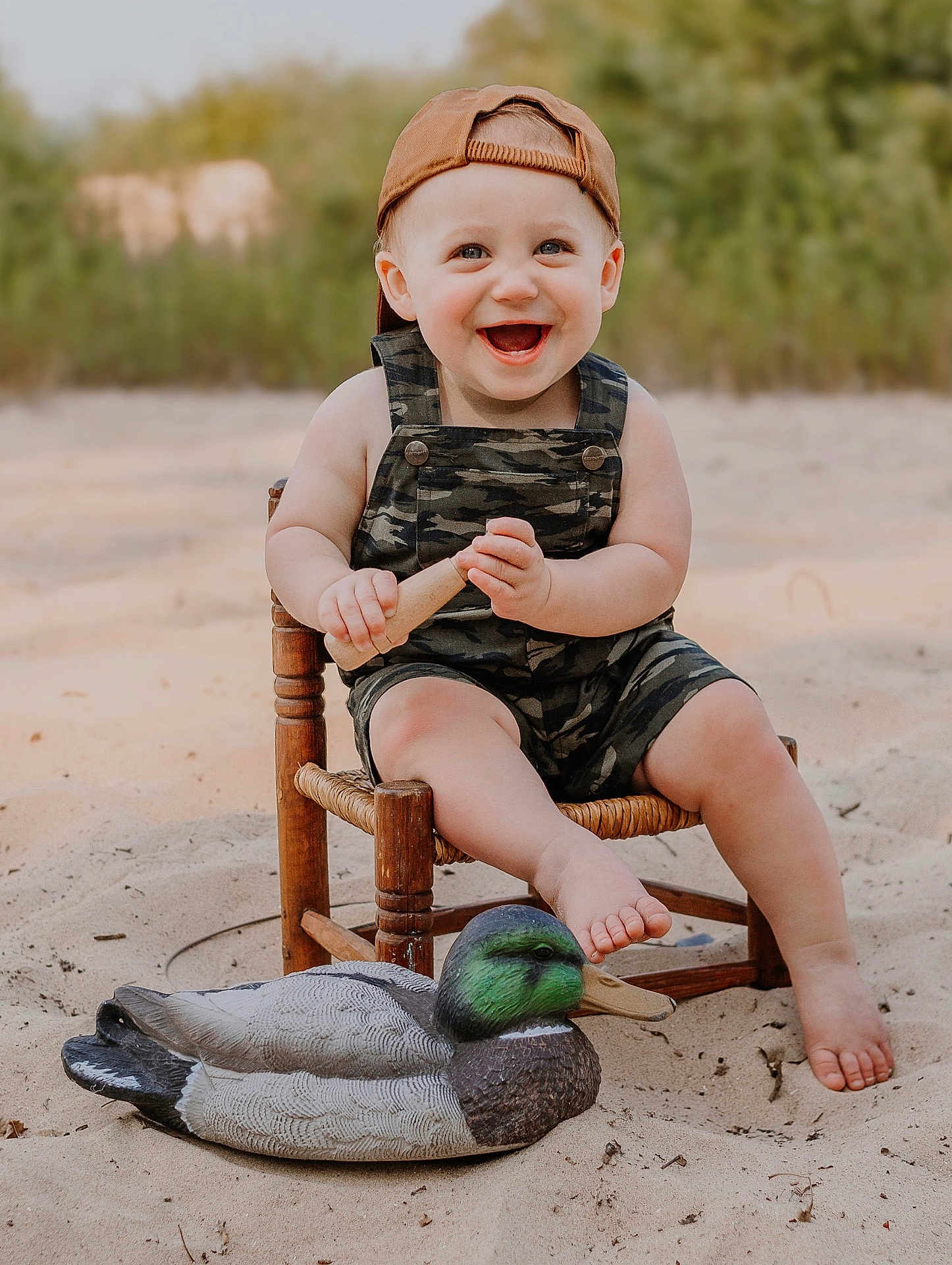 Nicholas is registered to the contest to win money with this photo: toddler, child, smiling, cap, brown_cap, overalls, camouflage, wooden_chair, sand, toy, wooden_toy, duck_decoy, outdoor, nature, greenery, happy, barefoot, portrait, seat, summer