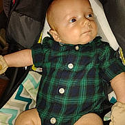 Timothy is registered to the contest to win money with this photo: baby, infant, plaid_clothing, onesie, mittens, cushion, baby_seat, fabric, pattern, comfort, child, skin, face, legs, arm, button, casual_wear, indoor, portrait, cute