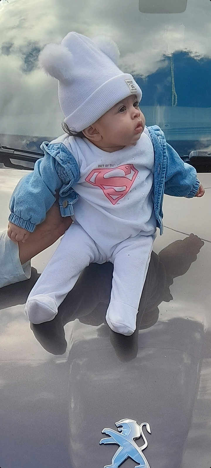 Aliyah a rejoint le concours — aidez-le/la à gagner de superbes lots ! baby, child, infant, hat, pom_pom, denim_jacket, superman_logo, white_clothing, car_hood, reflection, hand, outdoor, sky, clouds, portrait, cute, person, small_child, casual_clothing, fashion