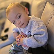 Theo is registered to the contest to win money with this photo: toddler, child, person, face, hands, couch, clothing, sweatshirt, pants, indoor, skin, expression, young_child, holding_hands, seated, looking_at_camera, soft_lighting, closeup, human, casual