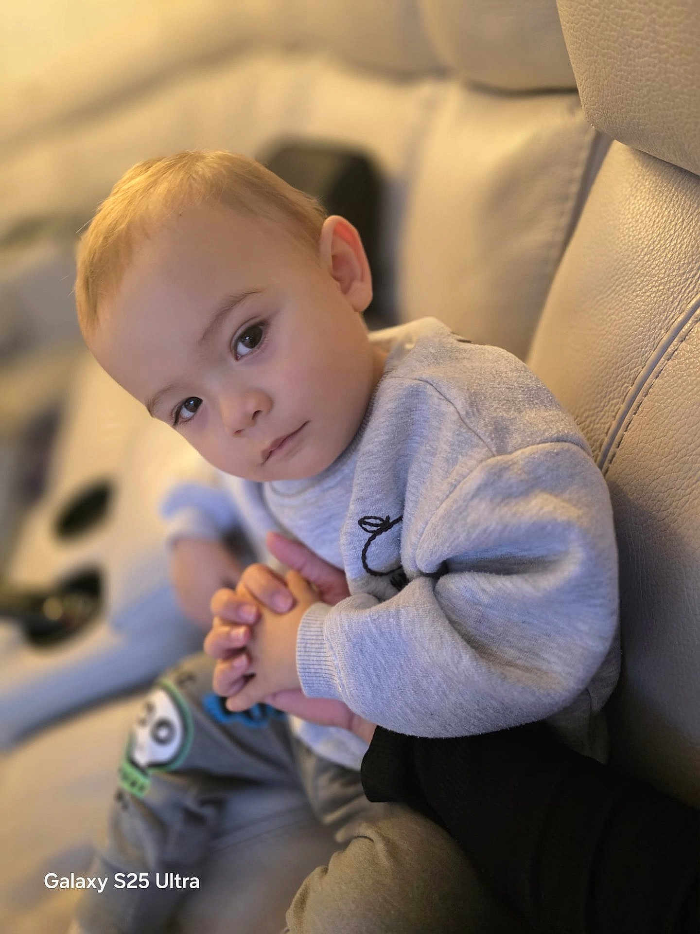 Theo is registered to the contest to win money with this photo: toddler, child, person, face, hands, couch, clothing, sweatshirt, pants, indoor, skin, expression, young_child, holding_hands, seated, looking_at_camera, soft_lighting, closeup, human, casual