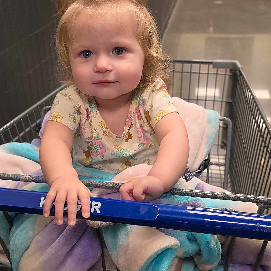 Delilah joined the competition — help win amazing prizes! baby, child, toddler, shopping_cart, grocery_store, indoor, portrait, sitting, serious_expression, blue_eyes, blonde_hair, ponytail, floral_shirt, blanket, barefoot, cart, aisle, hallway, fire_extinguisher, painting