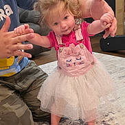 Delilah is registered to the contest to win money with this photo: child, toddler, girl, standing, smiling, pink_dress, tutu, overalls, barefoot, indoor, home, living_room, carpet, door, adult, hands, family, portrait, playful, dance