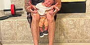 Cody Ryan is registered to the contest to win money with this photo: man, baby, child, person, indoor, carpet, fireplace, exercise_equipment, tattoo, cap, shorts, socks, smile, holding, knees, tiled_wall, casual_clothing, happy, bonding, home
