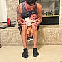man, baby, child, person, indoor, carpet, fireplace, exercise_equipment, tattoo, cap, shorts, socks, smile, holding, knees, tiled_wall, casual_clothing, happy, bonding, home